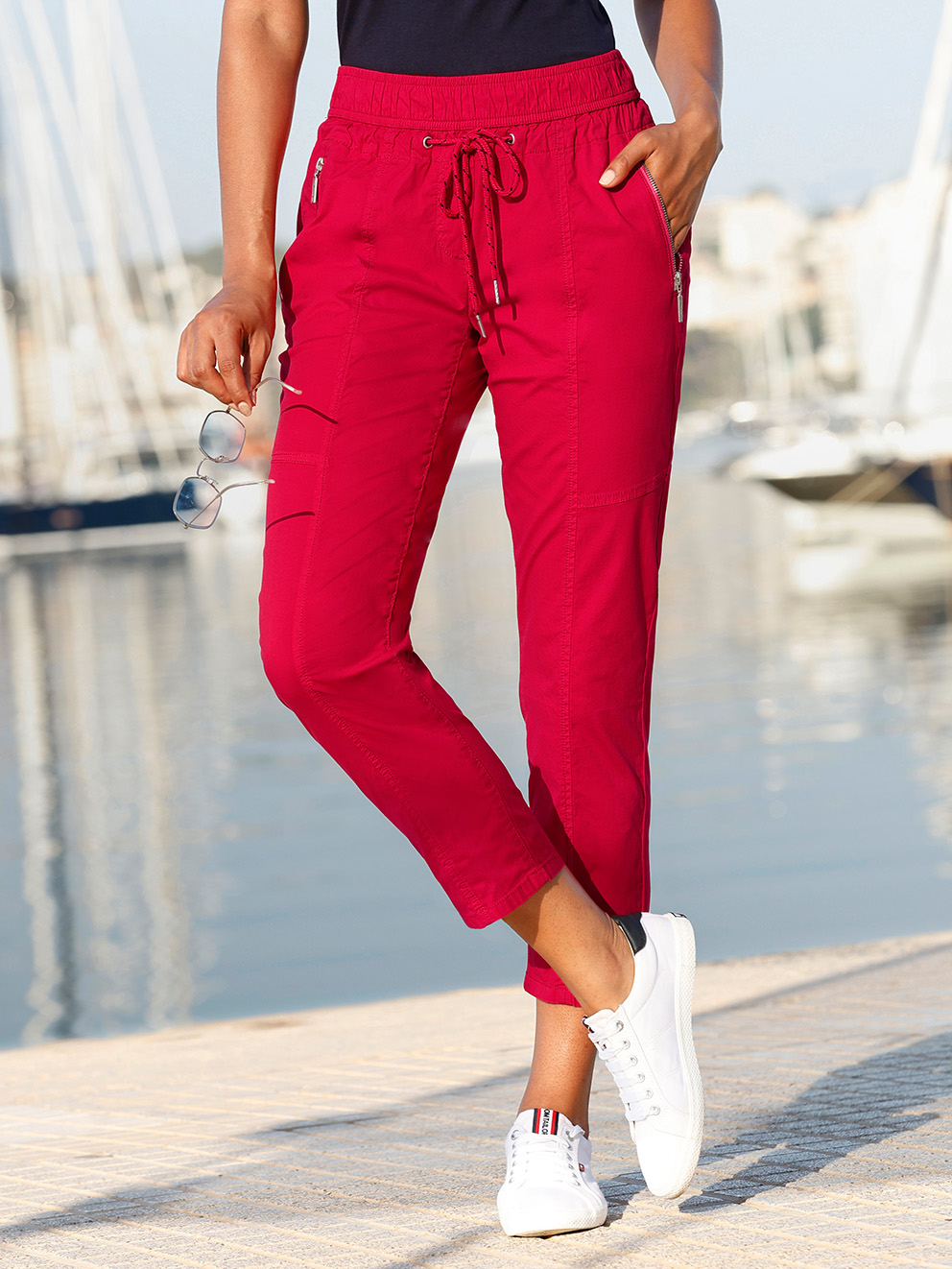 Ein Model steht am Hafen, trägt rote Jogpants mit Kordelzug, weiße Sneakers und hält eine Sonnenbrille in der Hand.