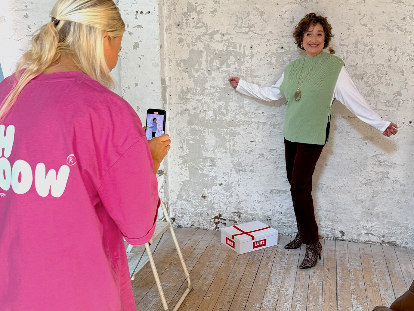 Eine Frau posiert vor einer weißen Wand, trägt einen grünen Pullunder, ein weißes Shirt, eine bordeauxrote Cordhose und Leo-Stiefeletten, eine Person fotografiert mit Smartphone.