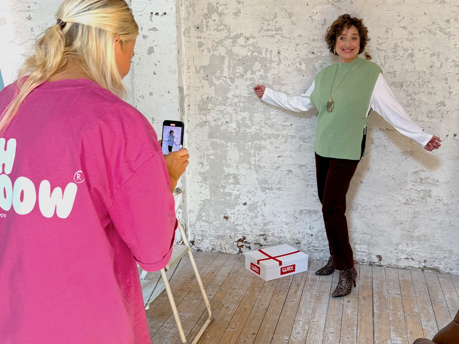 Eine Frau posiert vor einer weißen Wand, trägt einen grünen Pullunder, ein weißes Shirt, eine bordeauxrote Cordhose und Leo-Stiefeletten, eine Person fotografiert mit Smartphone. Eine Frau posiert vor einer weißen Wand, trägt einen grünen Pullunder, ein weißes Shirt, eine bordeauxrote Cordhose und Leo-Stiefeletten, eine Person fotografiert mit Smartphone.