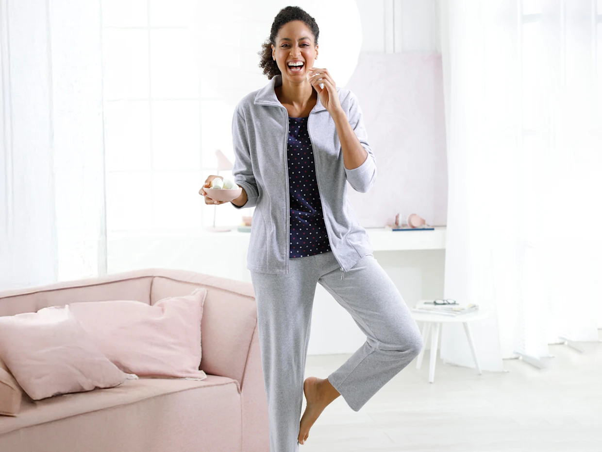 Ein Model steht barfuß in einem hellen Raum, trägt graue Freizeitkleidung und hält eine Tasse in der Hand.