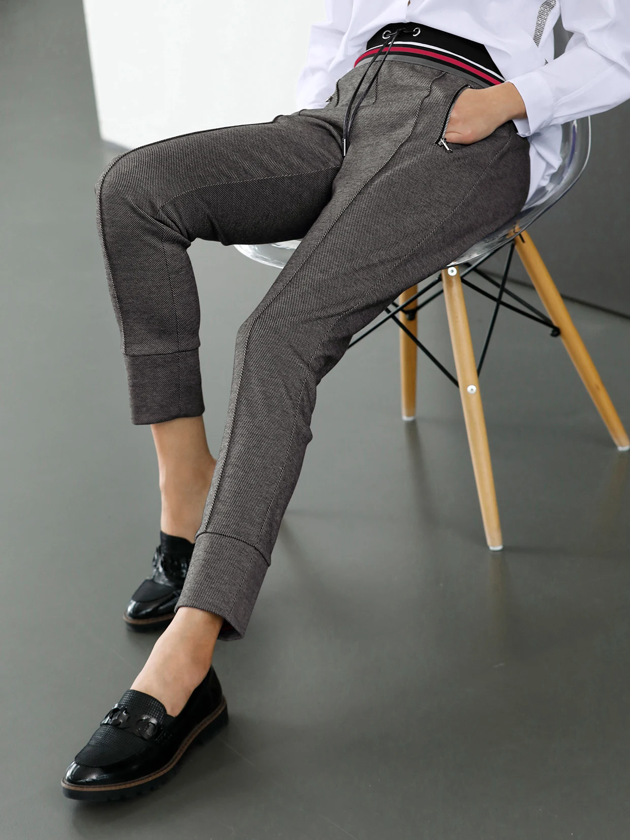 Grijze damesbroek met moderne pasvorm, gedragen met zwarte loafers en witte blouse, zittend op een transparante stoel.