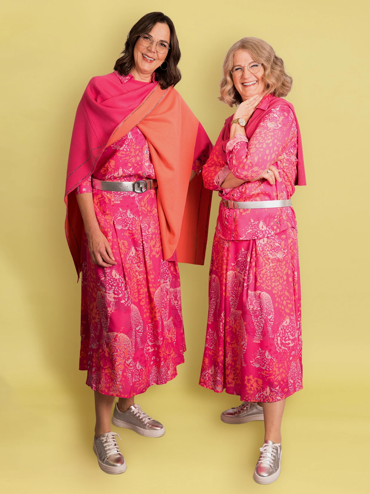 Zwei lächelnde Frauen stehen vor einer weißen Ziegelwand. Die Frau links hat dunkle Haare, eine Brille und trägt ein pink-orangefarbenes Tuch über einem pink gemusterten Kleid mit silbernem Gürtel und silbernen Sneakern. Die Frau rechts hat blonde Haare, eine Brille und trägt ebenfalls ein pink gemustertes Kleid mit einem pinken Blazer, silbernem Gürtel und silbernen Sneakern. Zwei lächelnde Frauen stehen vor einer weißen Ziegelwand. Die Frau links hat dunkle Haare, eine Brille und trägt ein pink-orangefarbenes Tuch über einem pink gemusterten Kleid mit silbernem Gürtel und silbernen Sneakern. Die Frau rechts hat blonde Haare, eine Brille und trägt ebenfalls ein pink gemustertes Kleid mit einem pinken Blazer, silbernem Gürtel und silbernen Sneakern.