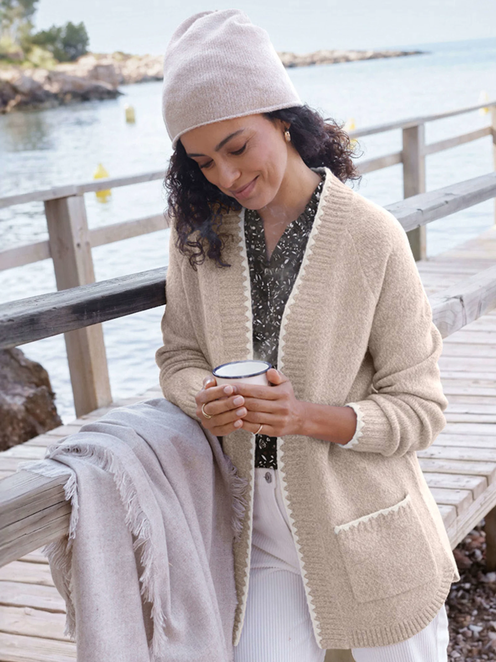 Ein Model mit Mütze und Strickjacke hält eine Tasse auf einem Holzsteg am Meer.