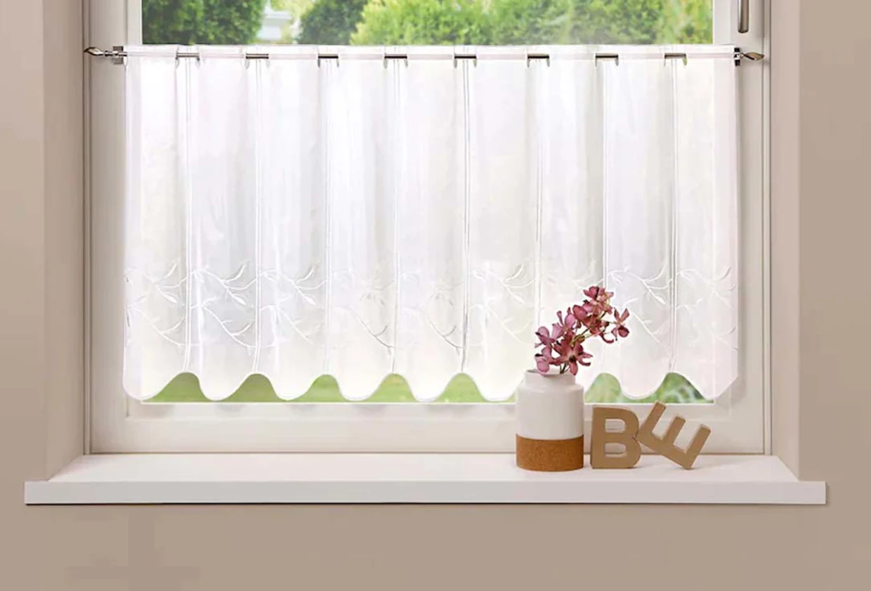 Halbtransparente weiße Scheibengardine mit zartem Muster, dekoriert mit einer Vase mit pinken Blüten und Buchstaben "BE" auf einer Fensterbank. Halbtransparente weiße Scheibengardine mit zartem Muster, dekoriert mit einer Vase mit pinken Blüten und Buchstaben "BE" auf einer Fensterbank.