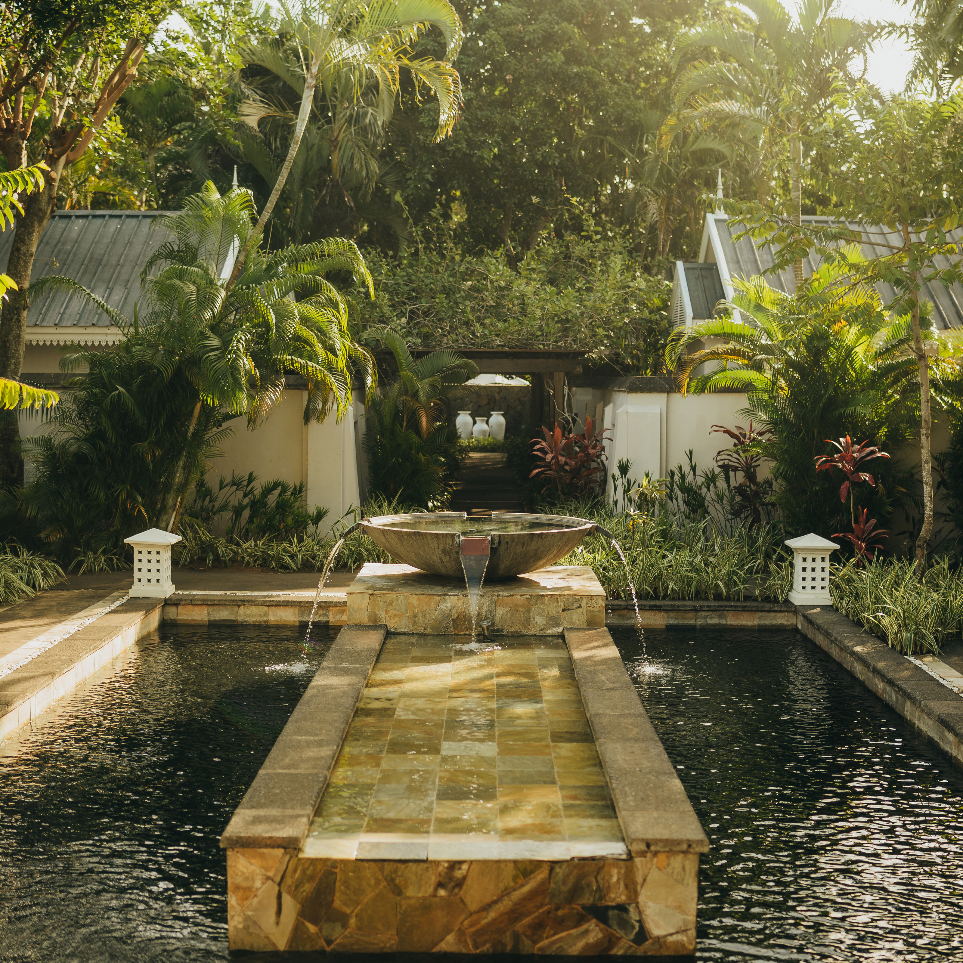 Ruhiger Garten mit Wasserbecken, kleinem Springbrunnen und tropischen Pflanzen, umgeben von Gebäuden.