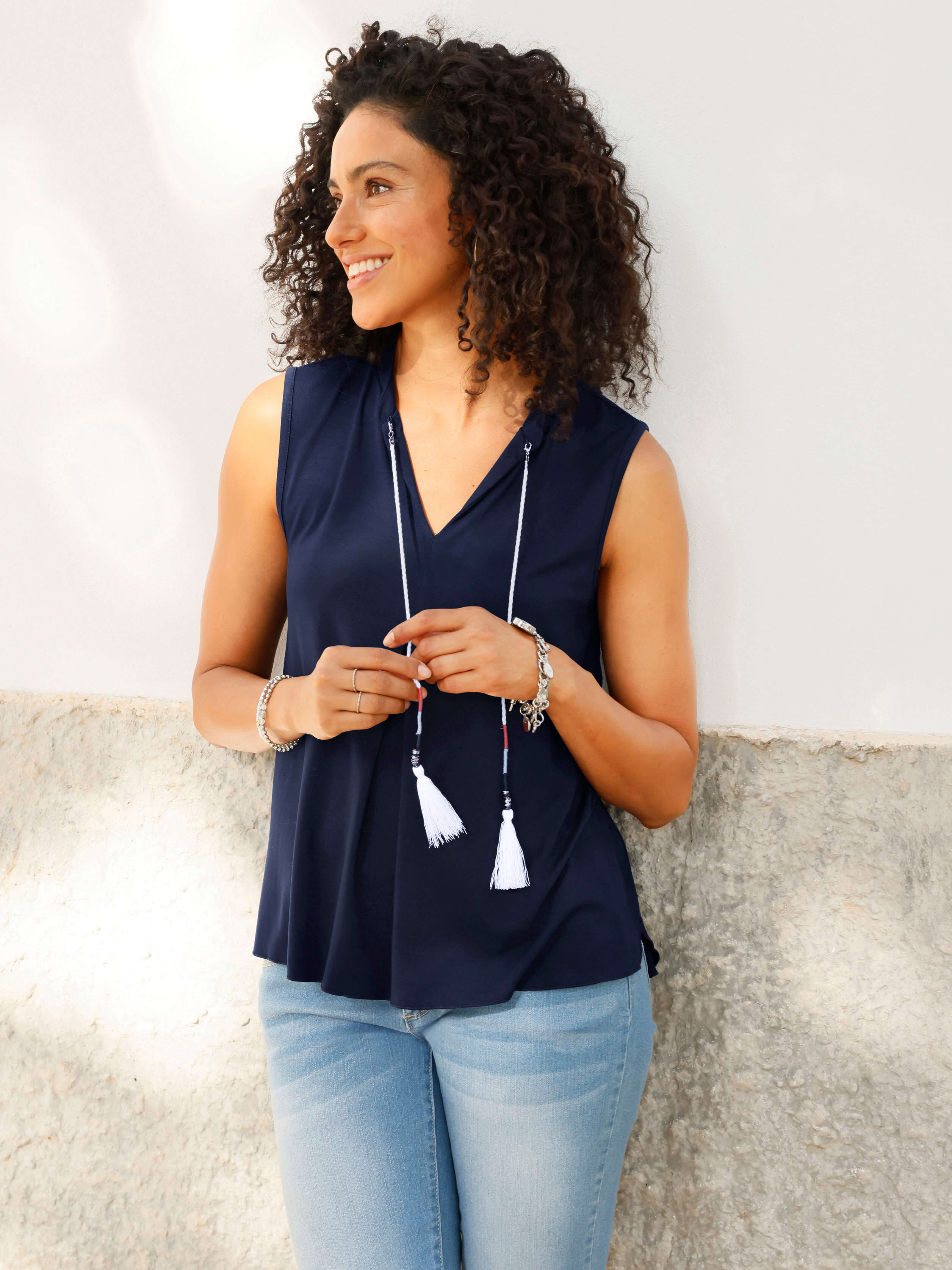 Ein Model steht an einer Wand, trägt eine ärmellose, dunkelblaue Bluse mit V-Ausschnitt, helle Jeans und eine lange Kette mit Quasten.