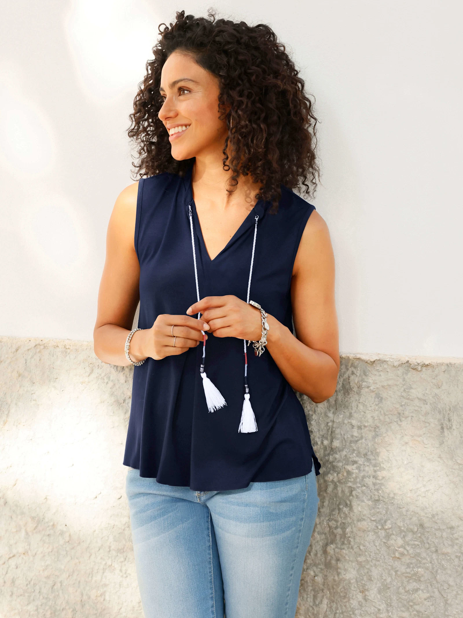 Ein Model steht an einer Wand, trägt eine ärmellose, dunkelblaue Bluse mit V-Ausschnitt, helle Jeans und eine lange Kette mit Quasten.