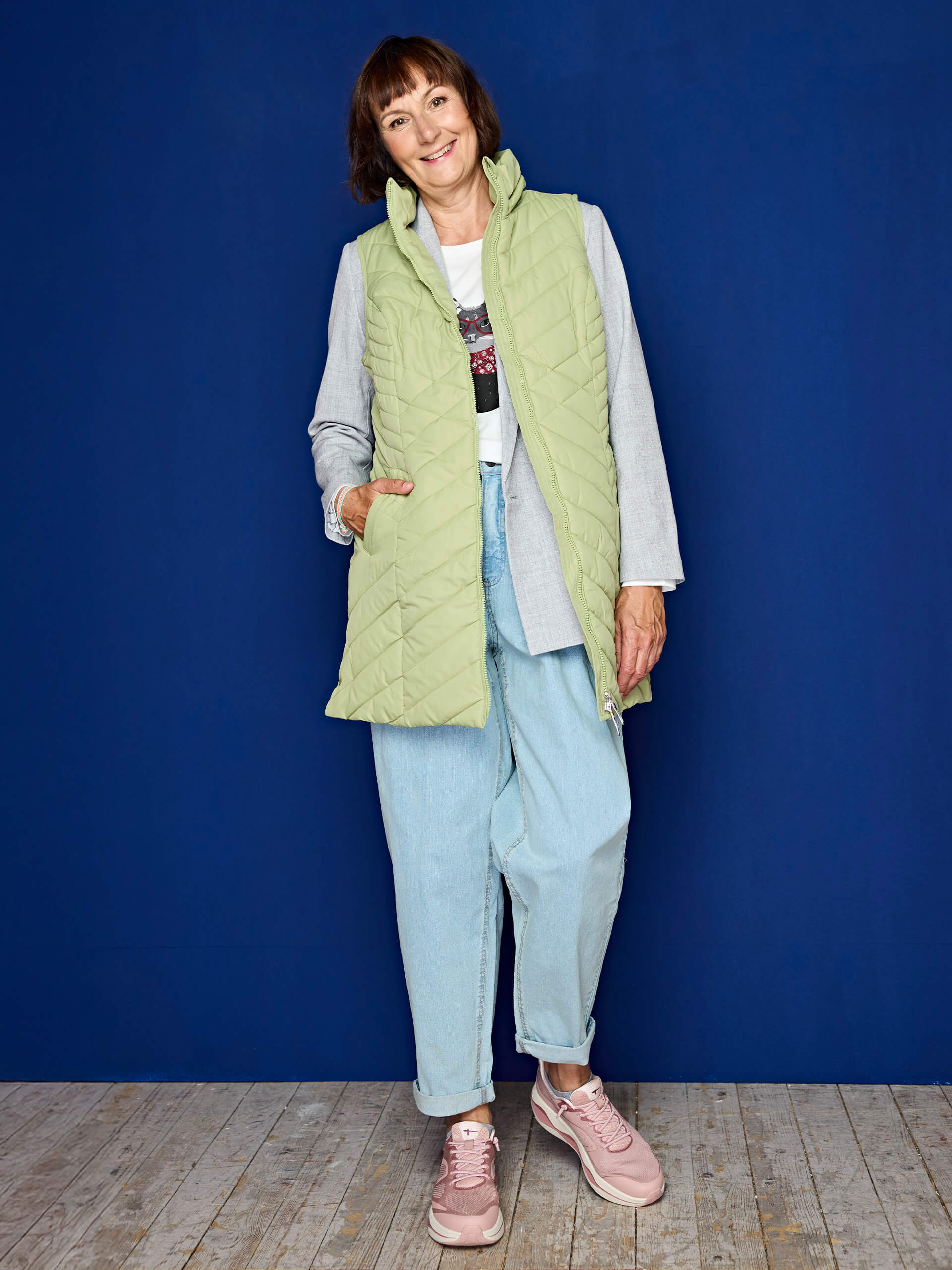 Eine Frau trägt eine hellgrüne gesteppte Weste, ein graues Sakko, ein weißes T-Shirt, helle Jeans und rosa Sneakers vor blauem Hintergrund.
