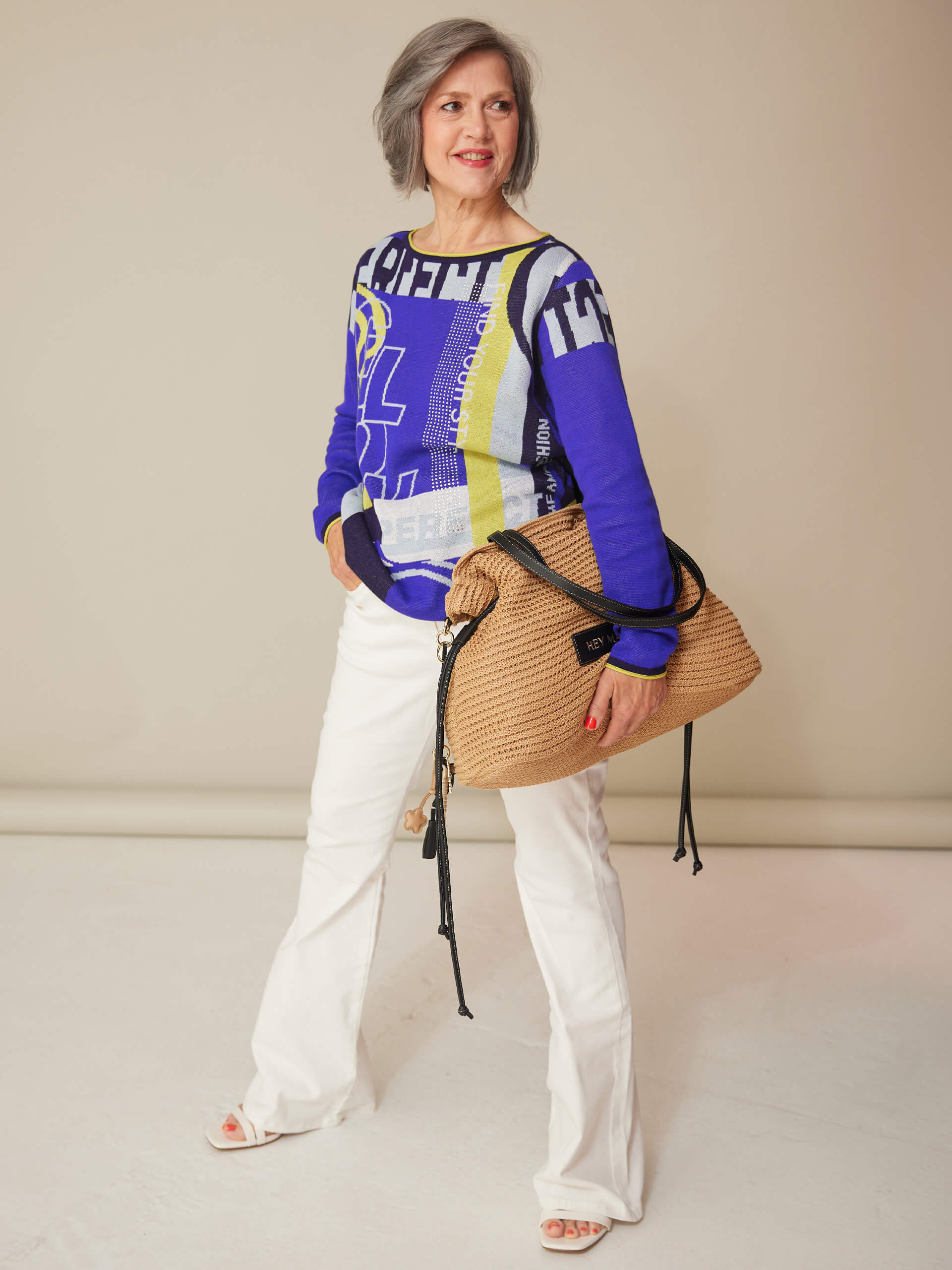 Ein Model trägt einen blau gemusterten Pullover, weiße Hose und Sandalen und hält eine große geflochtene Tasche vor beigefarbenem Hintergrund.