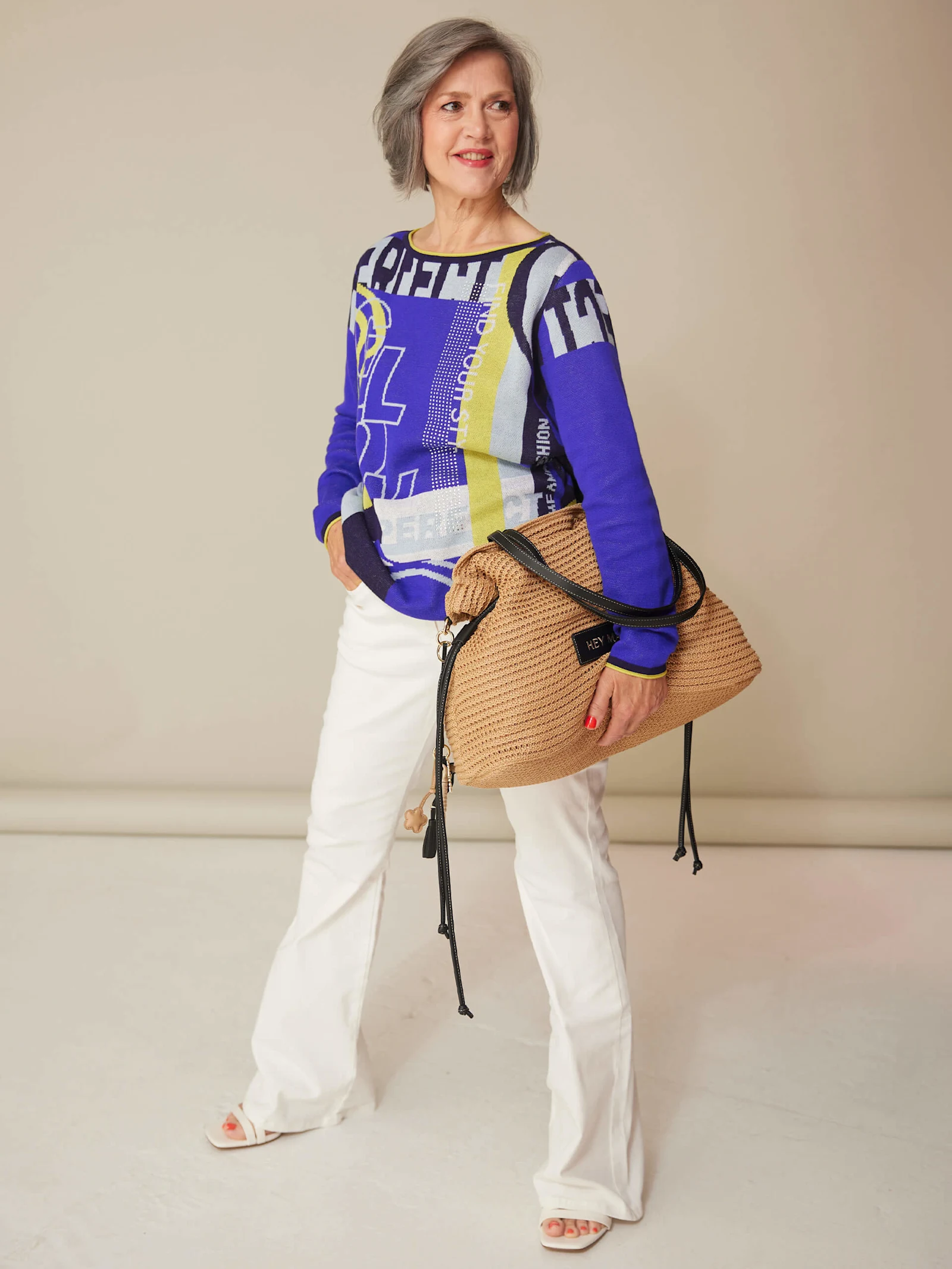 Ein Model trägt einen blau gemusterten Pullover, weiße Hose und Sandalen und hält eine große geflochtene Tasche vor beigefarbenem Hintergrund.