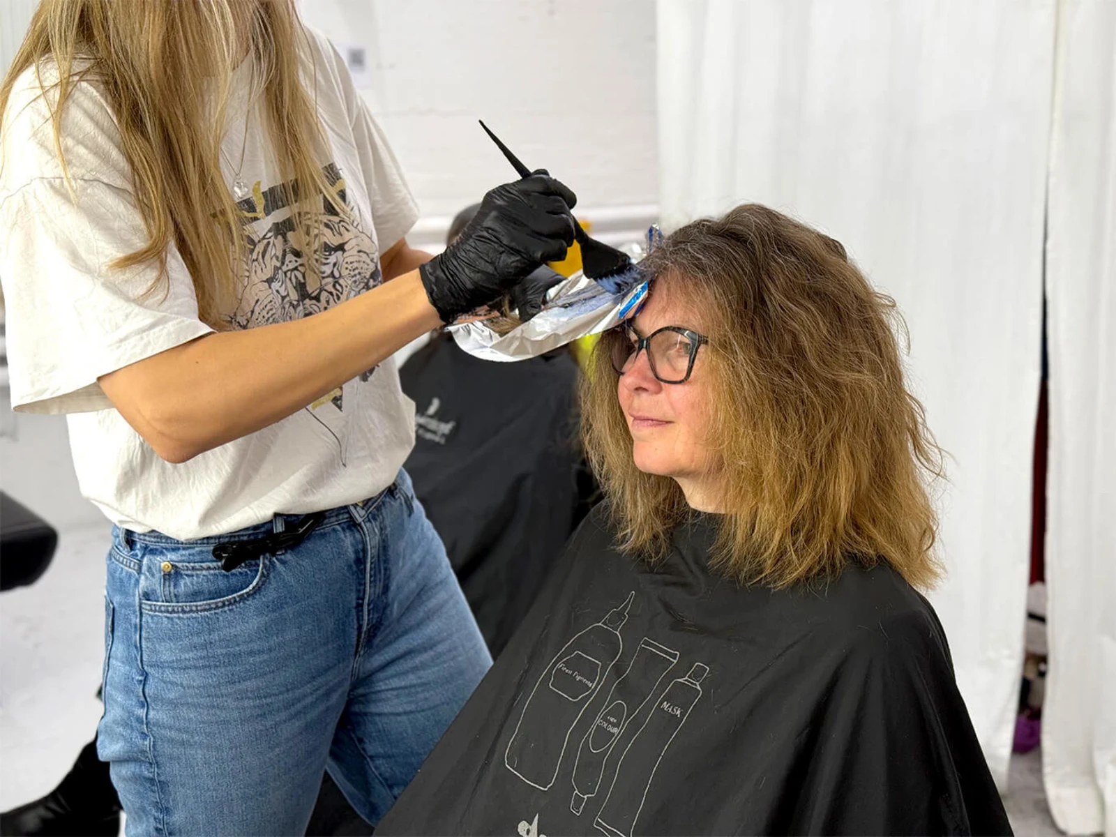 Eine Frau mit lockigem Haar sitzt in einem Friseurstuhl, während eine Person mit schwarzen Handschuhen Farbe auf das Haar aufträgt. Der Hintergrund ist unscharf. Eine Frau mit lockigem Haar sitzt in einem Friseurstuhl, während eine Person mit schwarzen Handschuhen Farbe auf das Haar aufträgt. Der Hintergrund ist unscharf.