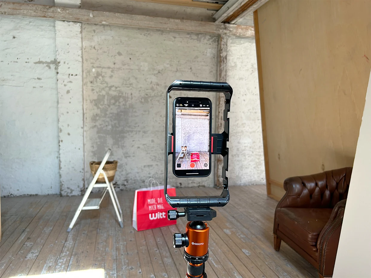 Smartphone auf Stativ in einem Raum mit Holzfußboden, braunem Ledersessel, weißer Leiter und roter Einkaufstasche im Hintergrund. Smartphone auf Stativ in einem Raum mit Holzfußboden, braunem Ledersessel, weißer Leiter und roter Einkaufstasche im Hintergrund.