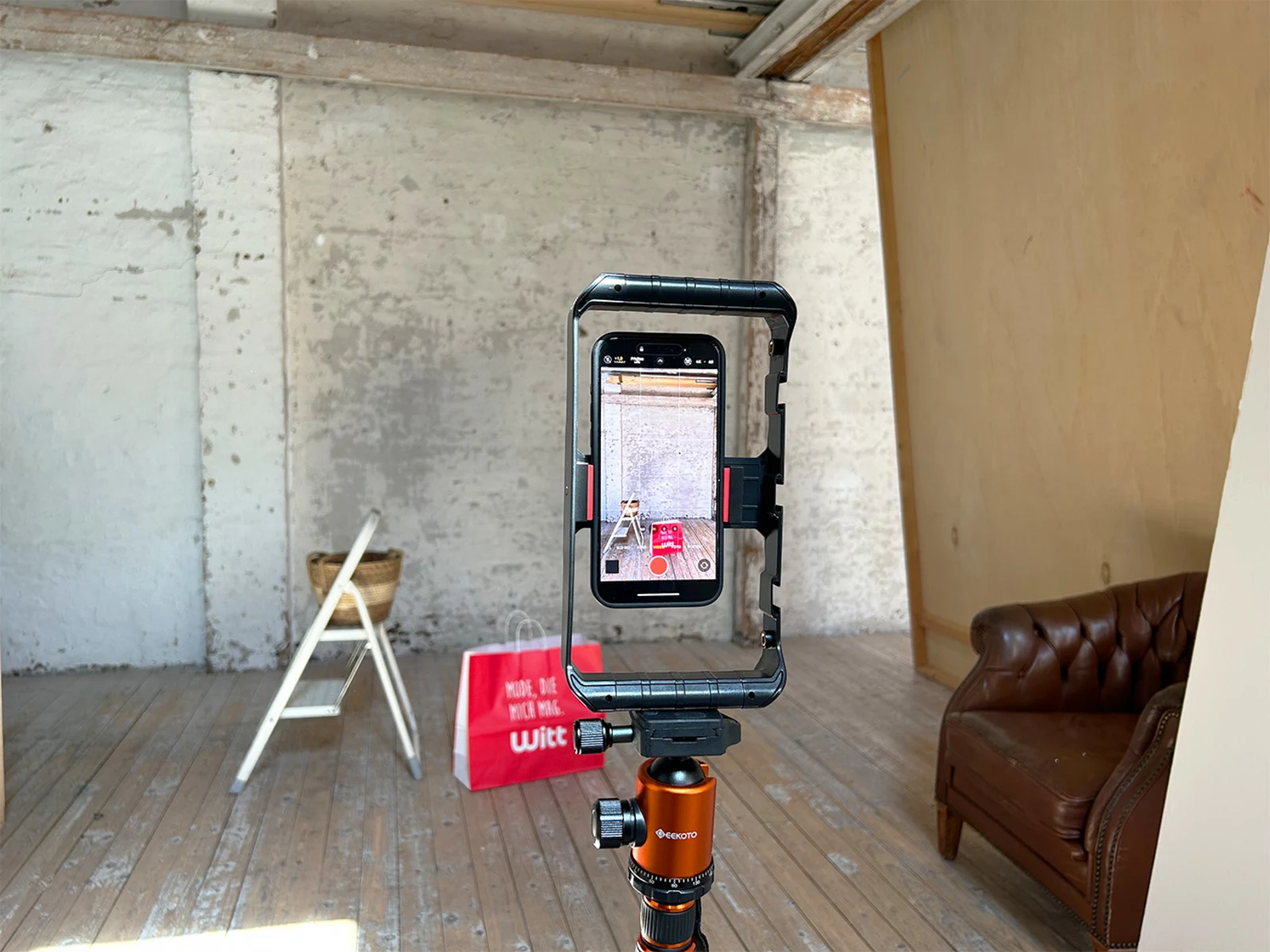Smartphone auf Stativ in einem Raum mit Holzfußboden, braunem Ledersessel, weißer Leiter und roter Einkaufstasche im Hintergrund. Smartphone auf Stativ in einem Raum mit Holzfußboden, braunem Ledersessel, weißer Leiter und roter Einkaufstasche im Hintergrund.