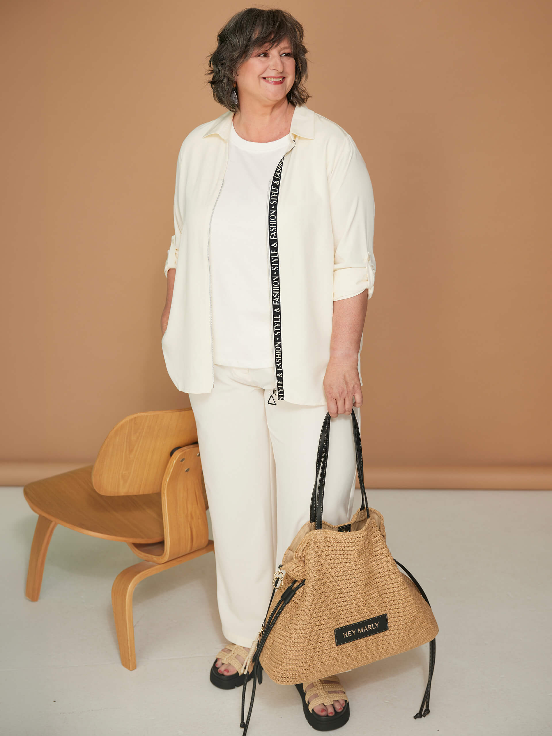 Ein Model trägt ein weißes Outfit und hält eine geflochtene Handtasche neben einem Holzstuhl.