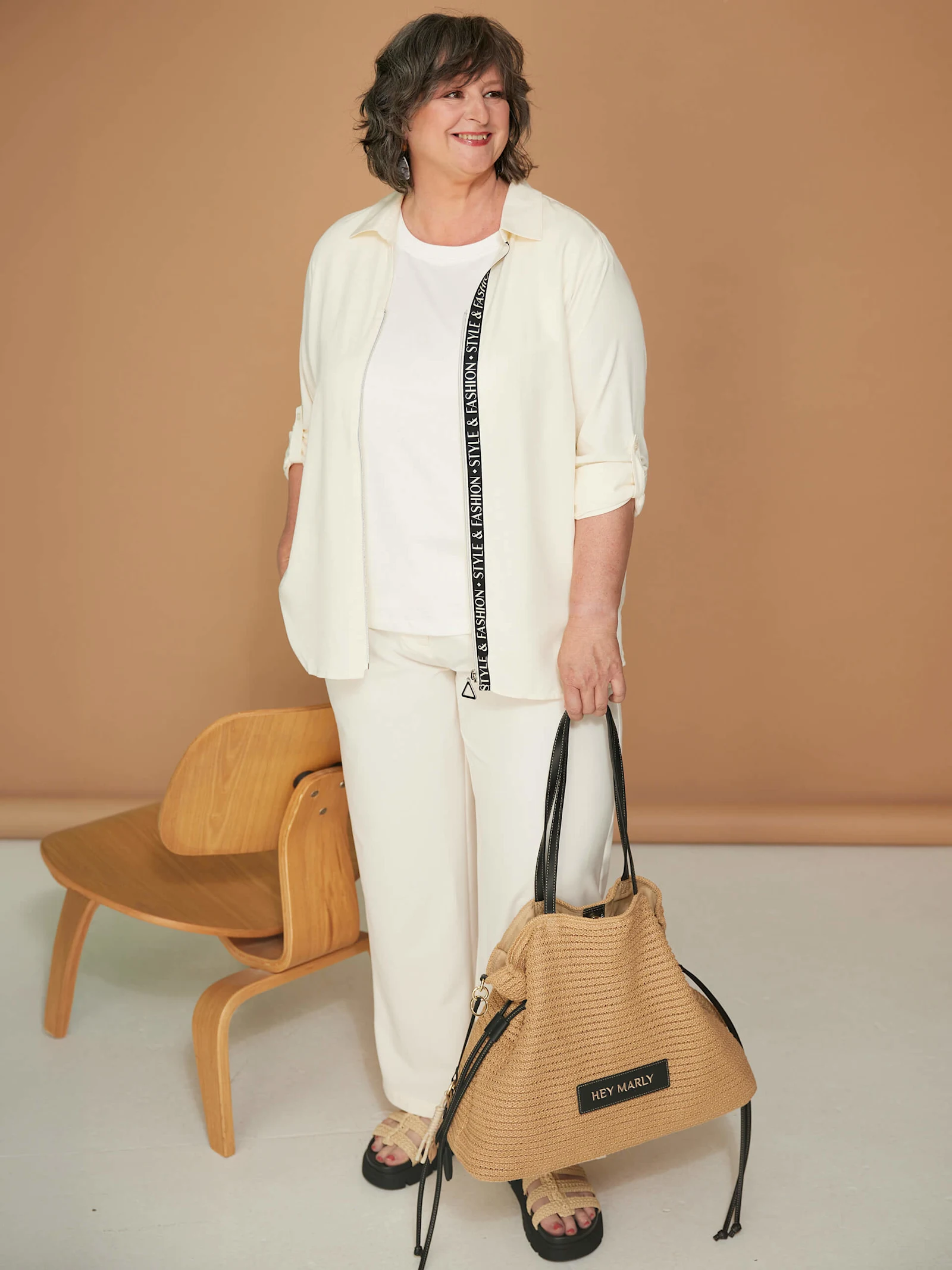 Ein Model trägt ein weißes Outfit und hält eine geflochtene Handtasche neben einem Holzstuhl.