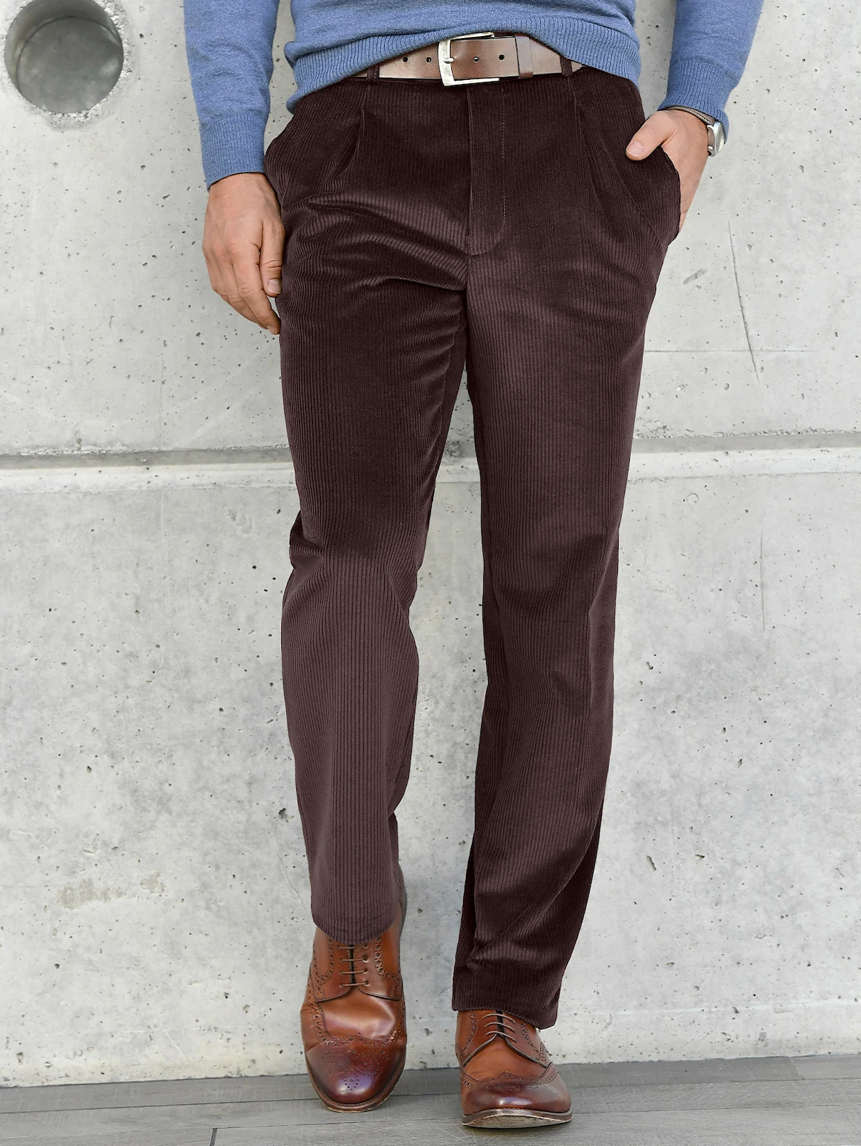 Braune Cordhose kombiniert mit einem blauen Pullover, braunem Ledergürtel und eleganten braunen Schnürschuhen vor einer grauen Betonwand. Braune Cordhose kombiniert mit einem blauen Pullover, braunem Ledergürtel und eleganten braunen Schnürschuhen vor einer grauen Betonwand.