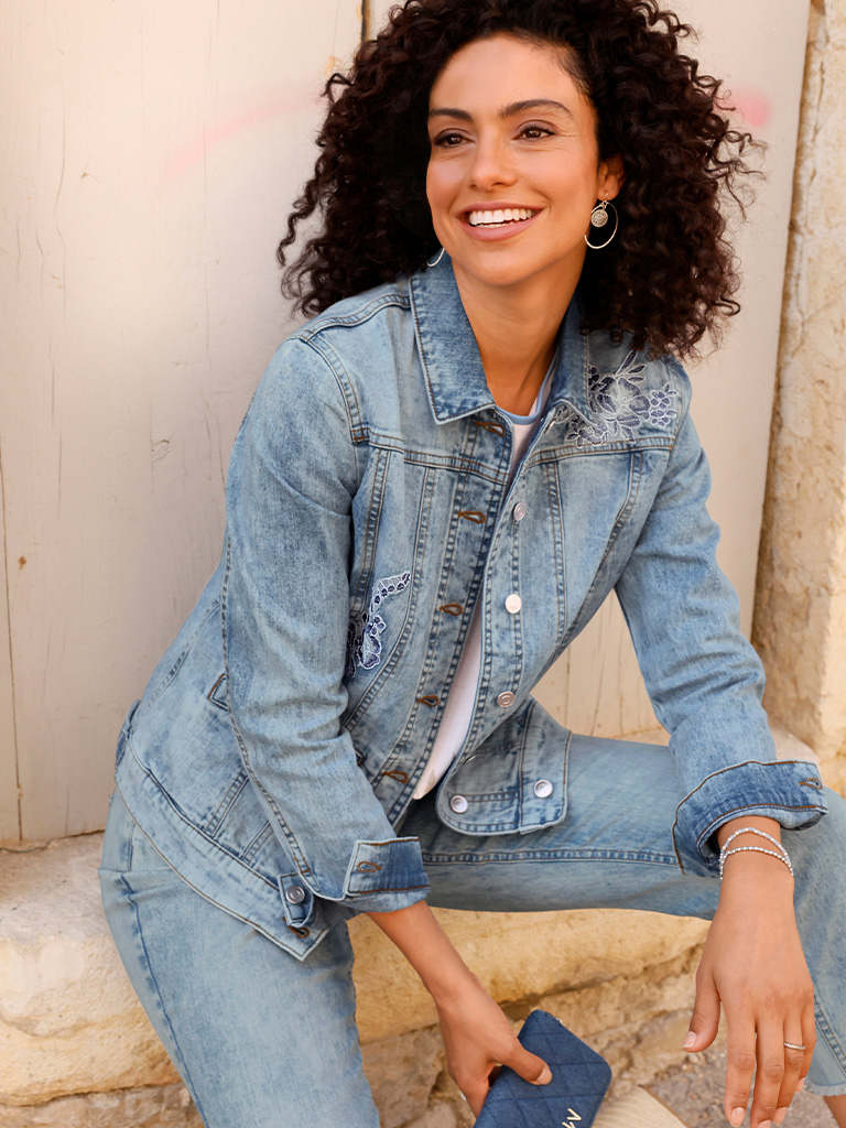 Ein Model trägt eine hellblaue Jeansjacke und eine hellblaue Jeanshose, sitzt im Freien und hält eine kleine blaue Tasche.