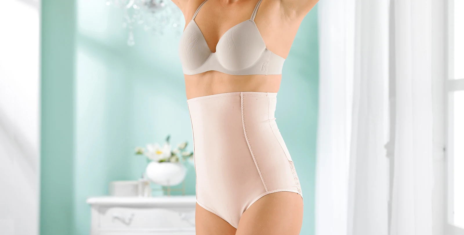 Frau trägt hautfarbenen figurformenden Mieder-Slip mit hoher Taille und einen passenden BH in heller Umgebung.