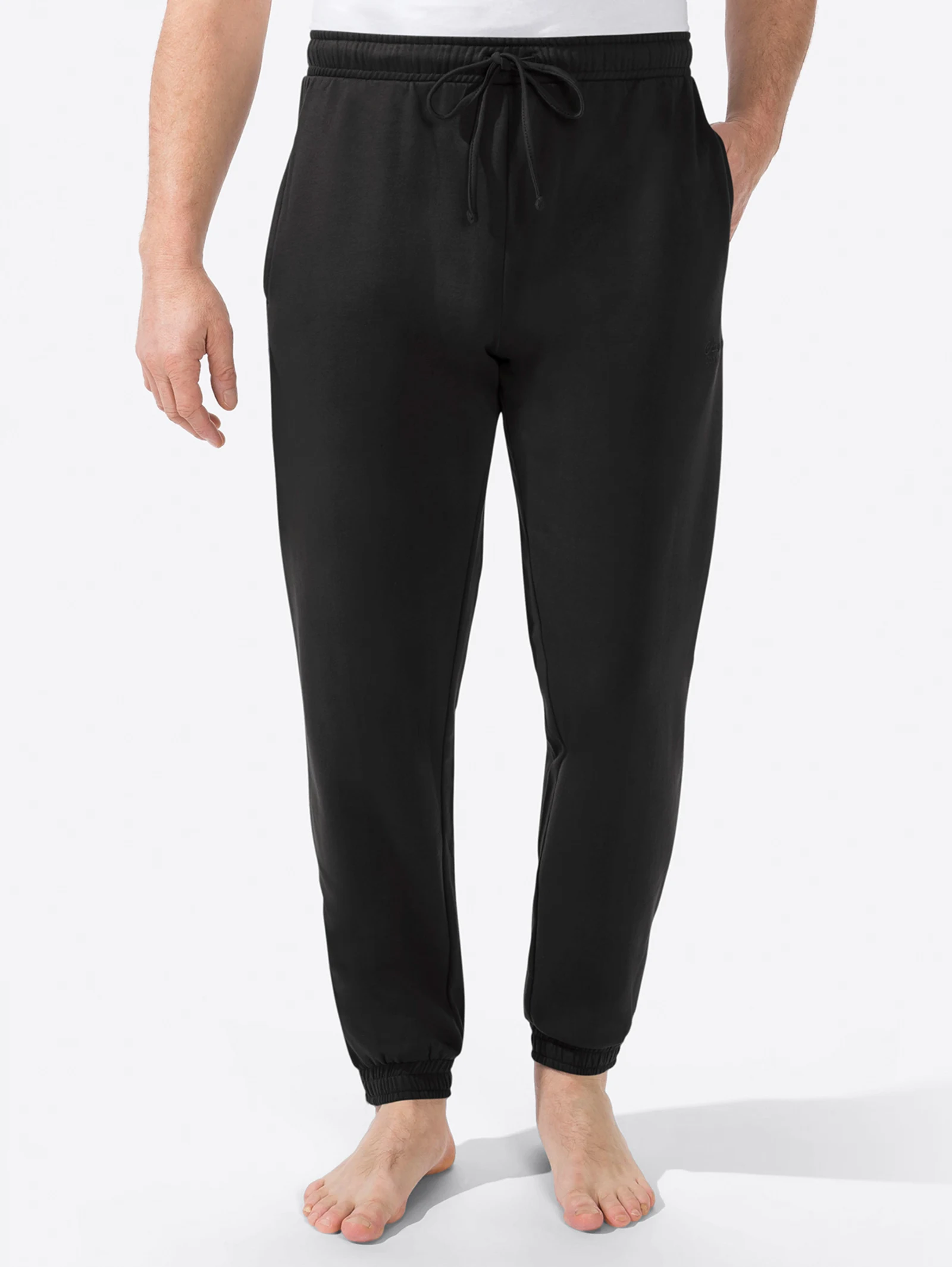 Schwarze Jogginghose für Herren mit elastischem Bund und Kordelzug, getragen von einer Person mit weißem T-Shirt, barfuß. Schwarze Jogginghose für Herren mit elastischem Bund und Kordelzug, getragen von einer Person mit weißem T-Shirt, barfuß.