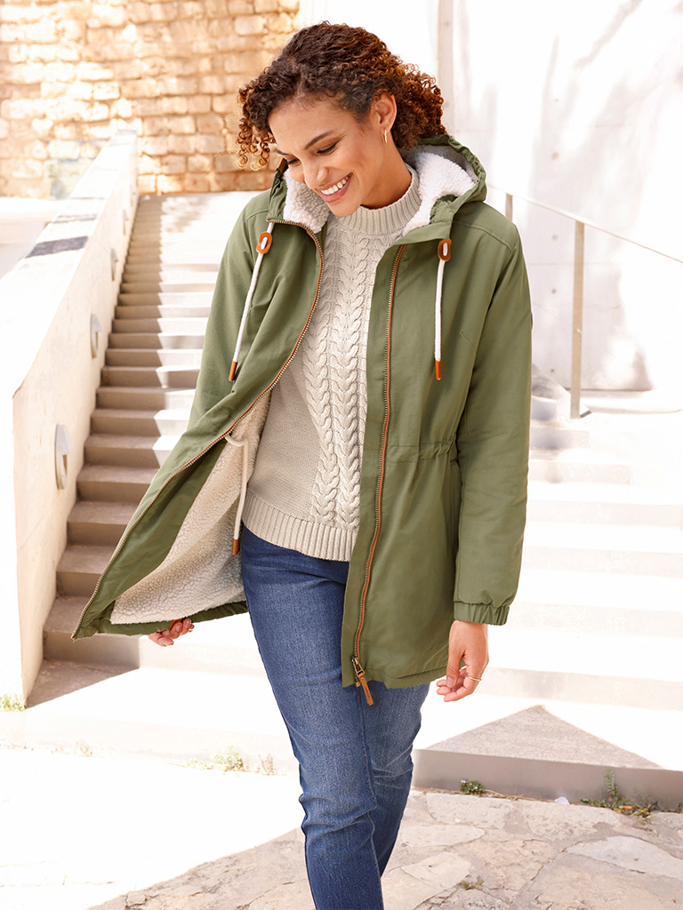 Ein Model trägt einen grünen, knielangen Parka mit Kapuze, darunter einen weißen, grob gestrickten Pullover und blaue Jeans, stehend im Freien.