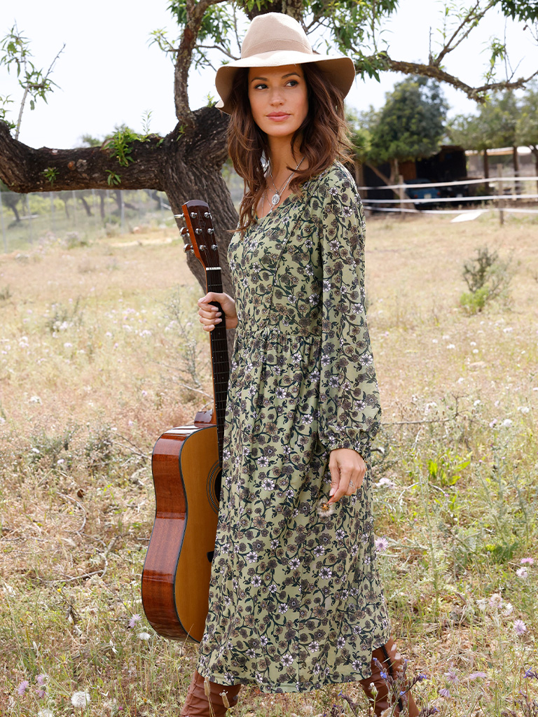 Ein Model trägt ein langes, schilf-bedrucktes Kleid und einen beigen Hut, steht auf einer Wiese und hält eine Gitarre.