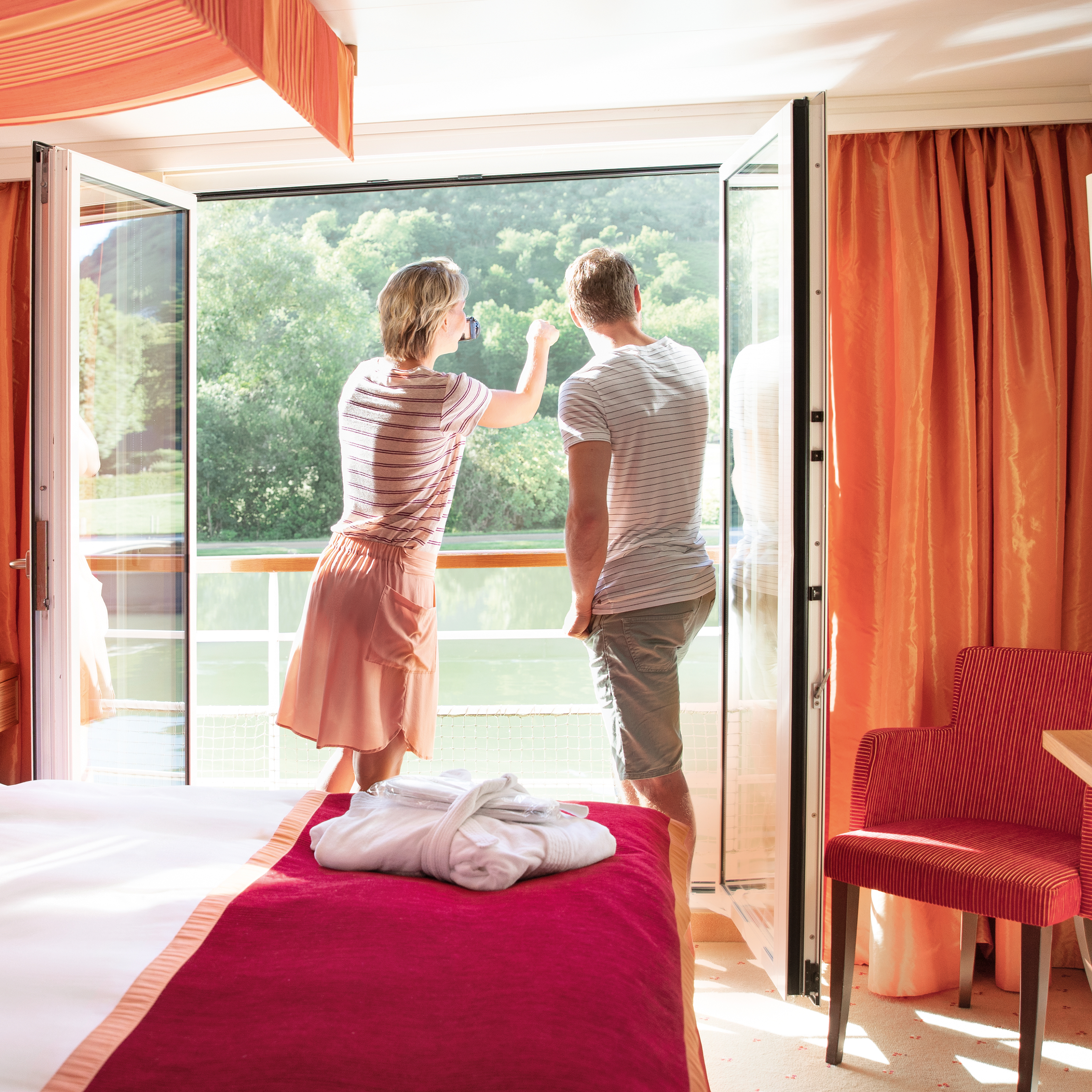 Ein Model steht in einem hellen Zimmer mit rotem Bett, blickt durch geöffnete Tür auf einen Fluss und grüne Natur.