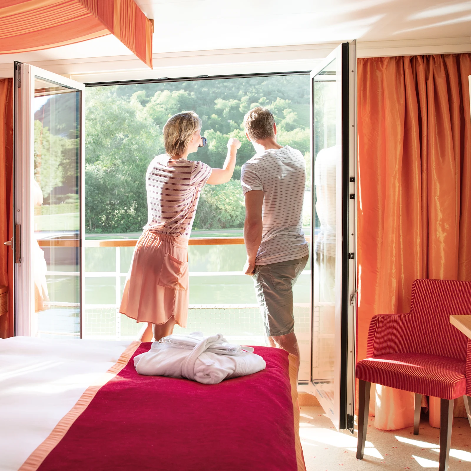 Ein Model steht in einem hellen Zimmer mit rotem Bett, blickt durch geöffnete Tür auf einen Fluss und grüne Natur. Ein Model steht in einem hellen Zimmer mit rotem Bett, blickt durch geöffnete Tür auf einen Fluss und grüne Natur.
