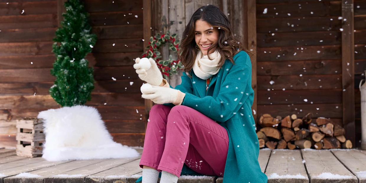 Een model zit buiten op een houten terras, draagt een groene jas, roze broek en witte handschoenen, omringd door vallende sneeuw.