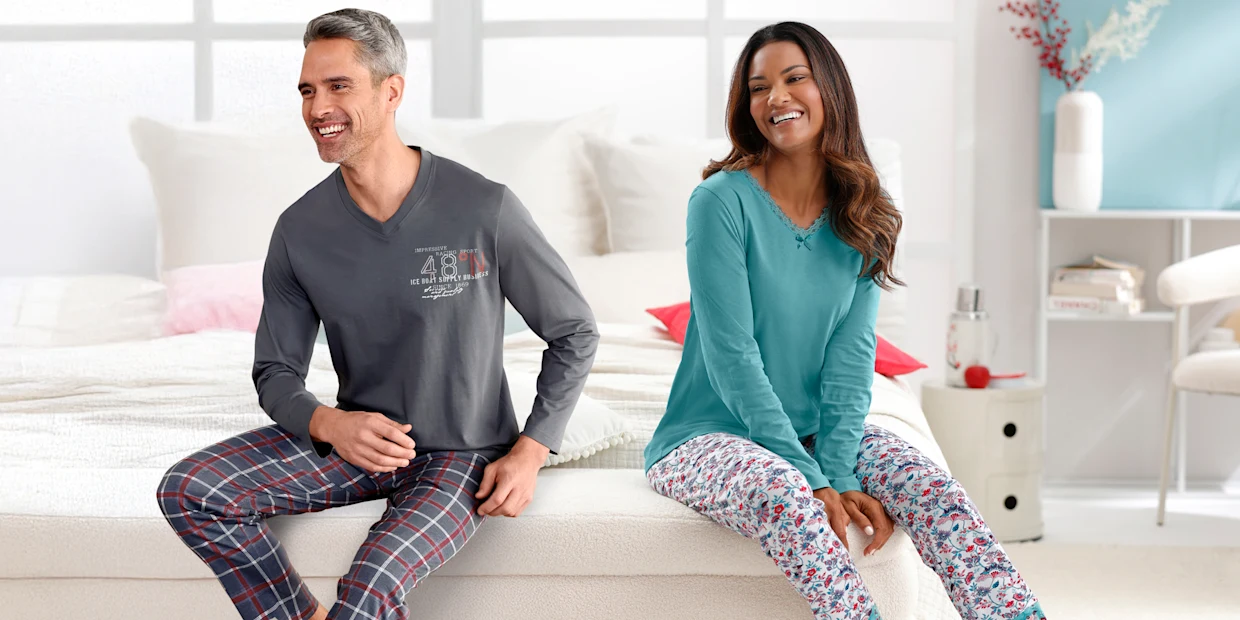Een mannelijk model met een grijs shirt met lange mouwen en geruite pyjamabroek zit naast een vrouwelijk model met een turquoise shirt met lange mouwen en een broek met patroon op een bed.