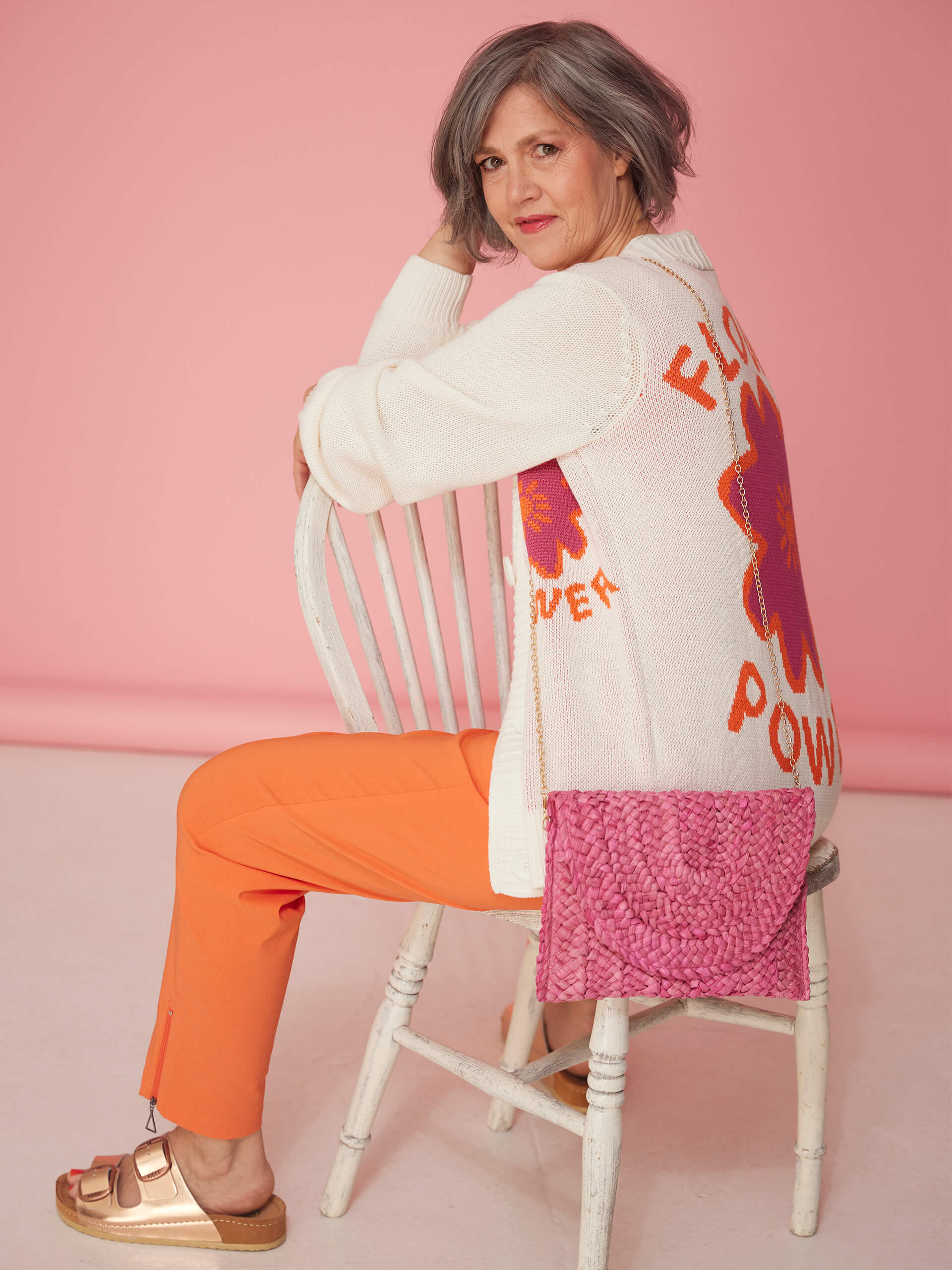 Ein Model sitzt auf einem weißen Stuhl und trägt eine cremefarbene Strickjacke, rosa Top, orange Hose und goldfarbene Sandalen.