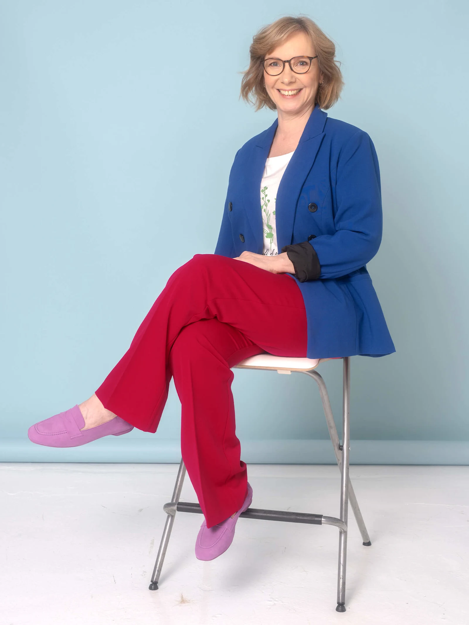 Eine Frau sitzt auf einem Stuhl, trägt einen blauen Blazer über einem weißen T-Shirt mit Printaufdruck, eine rote Hose und rosa Schuhe.