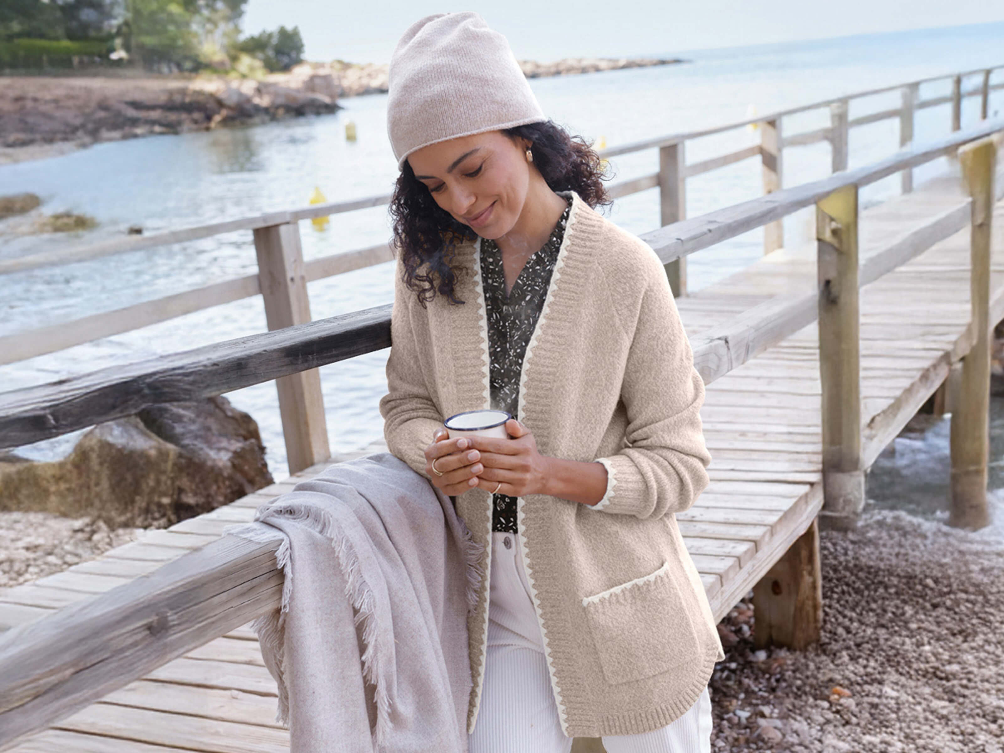 Ein Model mit Mütze und Strickjacke hält eine Tasse auf einem Holzsteg am Meer.