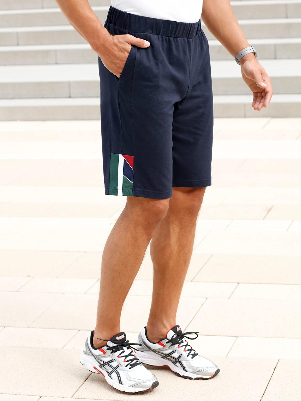 Mann trägt dunkelblaue Shorts mit seitlichem Farbdetail und weiße Sportschuhe, stehend auf hellen Pflastersteinen. Mann trägt dunkelblaue Shorts mit seitlichem Farbdetail und weiße Sportschuhe, stehend auf hellen Pflastersteinen.