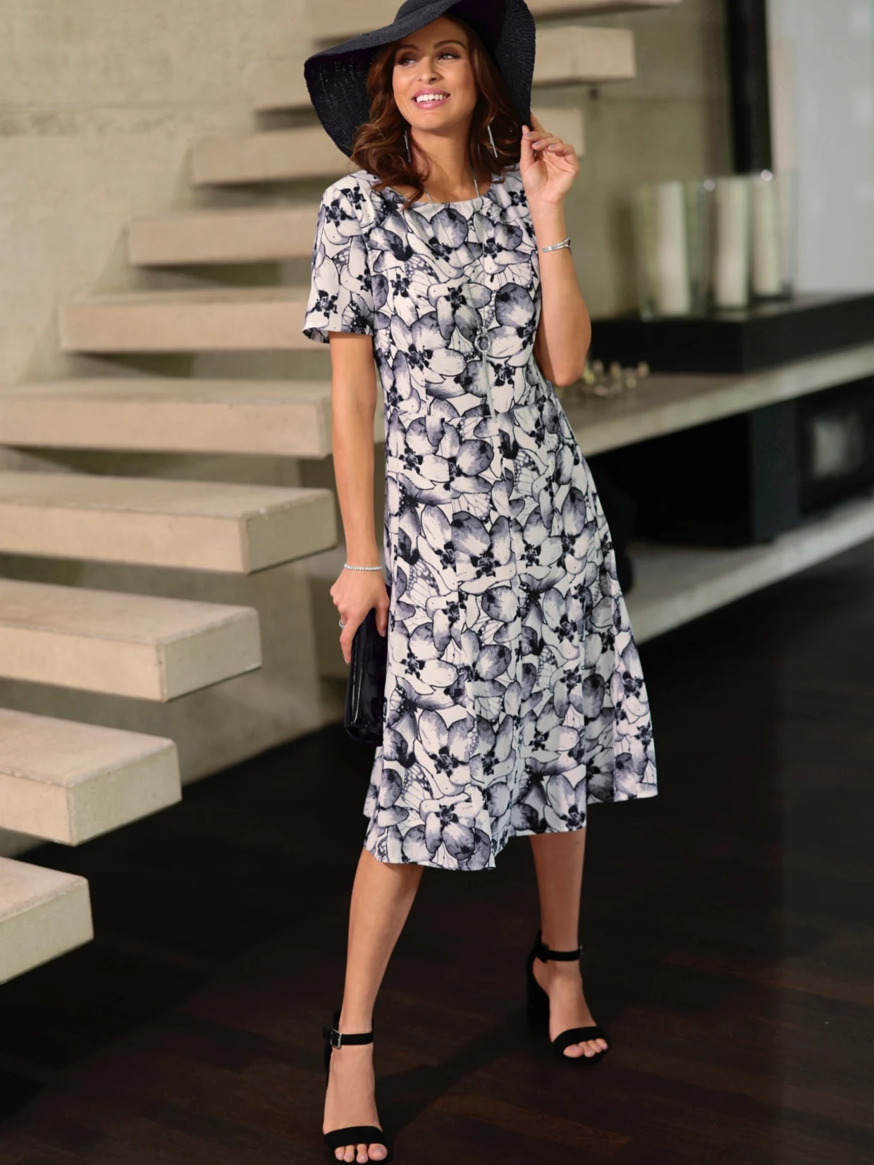 Frau trägt ein florales, knielanges Kleid in Schwarz-Weiß, kombiniert mit schwarzen Sandaletten, einem schwarzen Hut und einer kleinen Clutch. Im Hintergrund moderne Treppe und Dekoration.