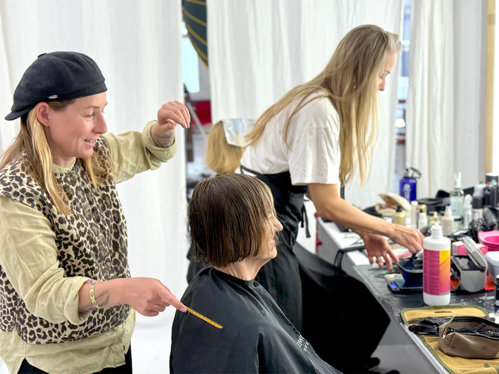 Auf dem Bild sind drei Frauen zu sehen. Eine Frau schneidet einer anderen mit schwarzem Friseurkittel, die vor ihr auf einem Stuhl sitzt, die Haare, während die dritte Frau im Hintergrund Produkte auf einem Tisch ordnet. Auf dem Bild sind drei Frauen zu sehen. Eine Frau schneidet einer anderen mit schwarzem Friseurkittel, die vor ihr auf einem Stuhl sitzt, die Haare, während die dritte Frau im Hintergrund Produkte auf einem Tisch ordnet.