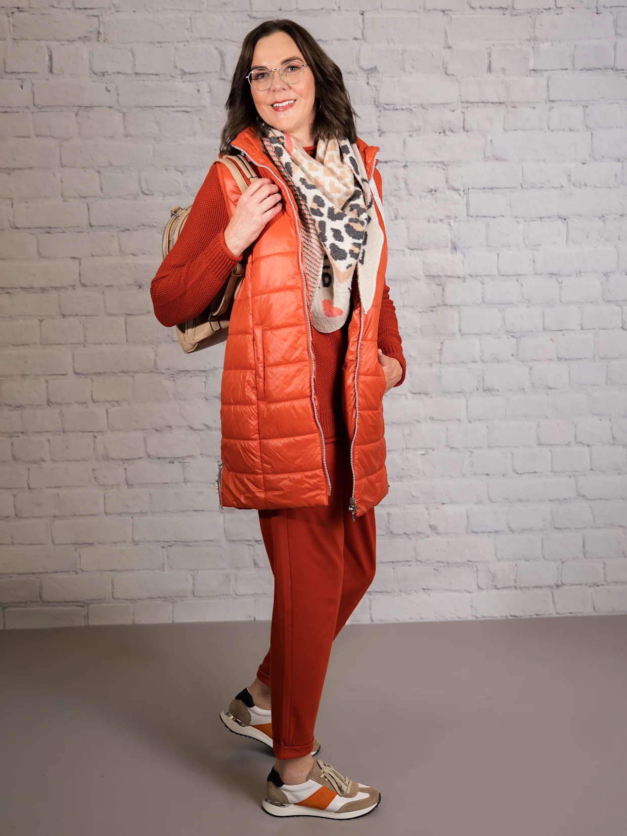 Eine dunkelhaarige Frau trägt eine rostrote Longweste, einen rostroten Pullover, rostrote Jogg Pants in Interlock-Qualität, einen beigen Dreieckschal und weiß-orange Sneaker. Der Hintergrund ist eine weiße Wand.
