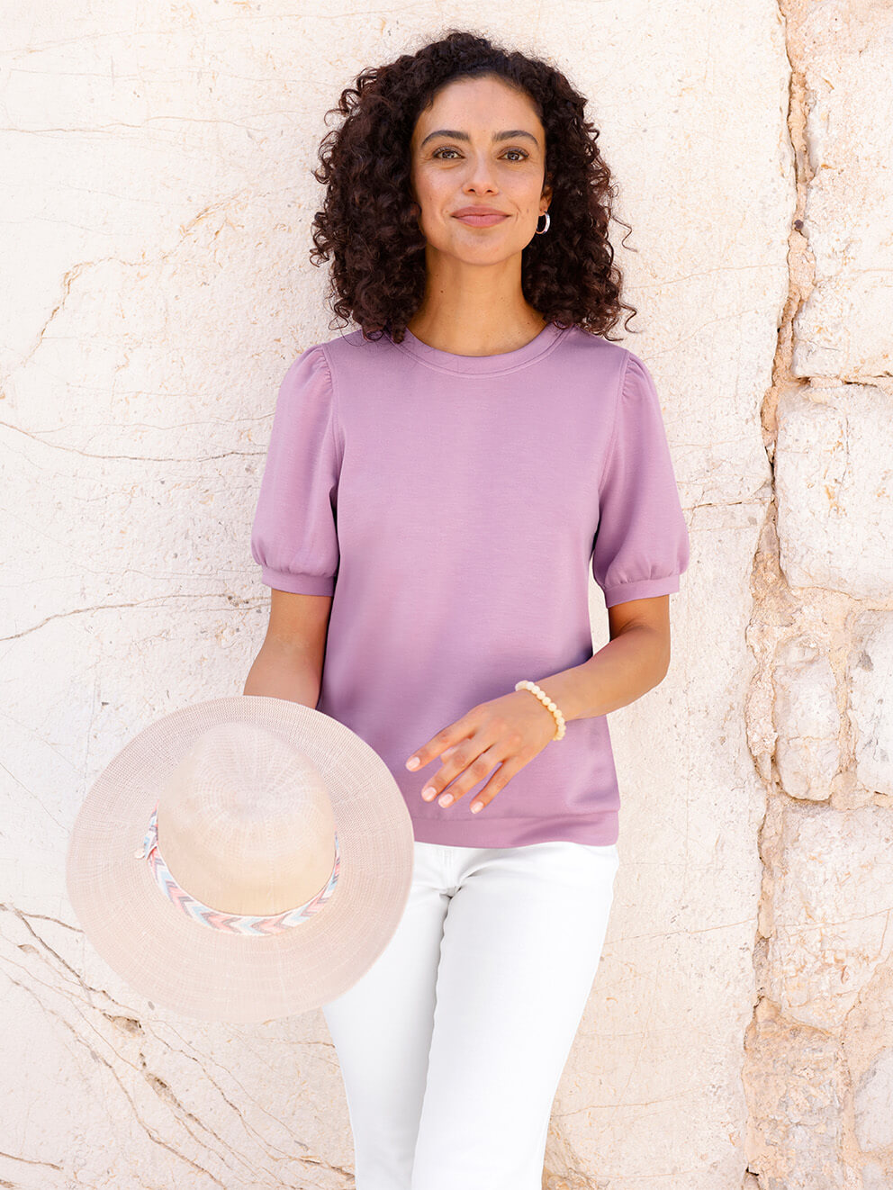 Ein Model trägt ein hellviolettes Shirt mit kurzen Puffärmeln und eine weiße Hose, hält einen beigen Sonnenhut, steht vor einer hellen Steinmauer.