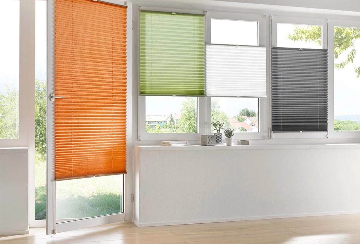 Vier moderne Plissees in den Farben Orange, Grün, Weiß und Anthrazit an bodentiefen Fenstern in einem hellen Raum. Vier moderne Plissees in den Farben Orange, Grün, Weiß und Anthrazit an bodentiefen Fenstern in einem hellen Raum.