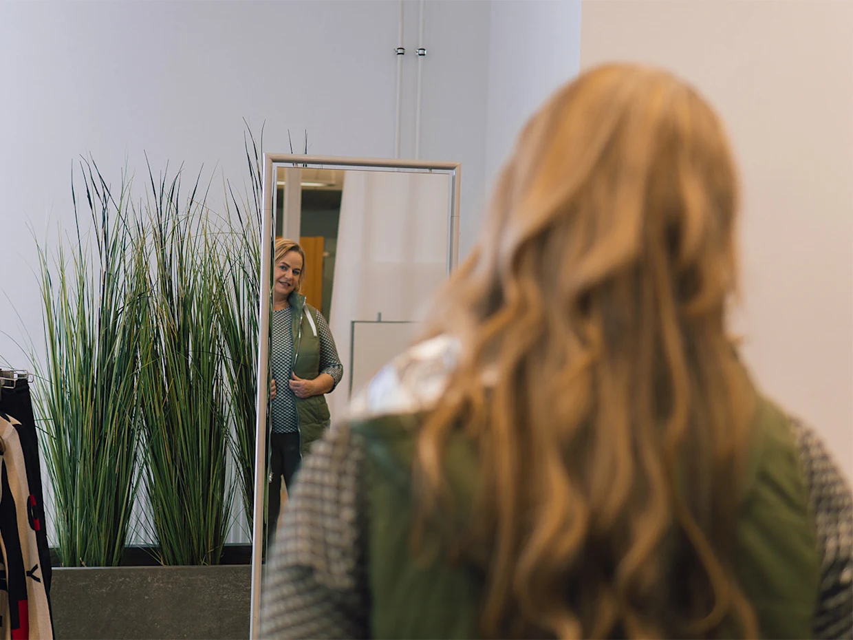 Eine blonde Frau steht mit dem Rücken zur Kamera und betrachtet sich in einem großen Spiegel. Im Hintergrund stehen grüne Topfpflanzen, der Raum ist hell mit weißen Wänden. Eine blonde Frau steht mit dem Rücken zur Kamera und betrachtet sich in einem großen Spiegel. Im Hintergrund stehen grüne Topfpflanzen, der Raum ist hell mit weißen Wänden.