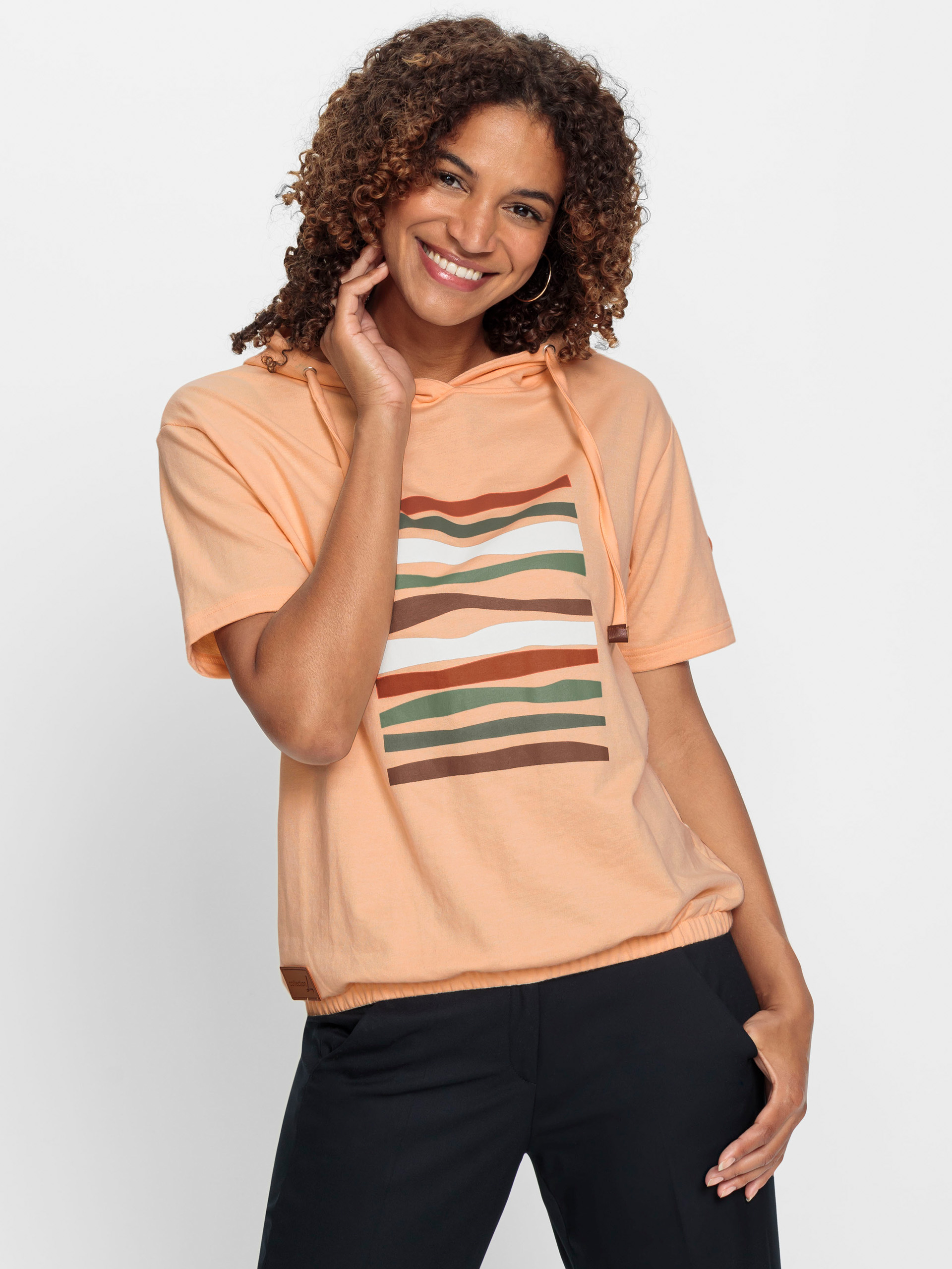 Frau trägt ein pfirsichfarbenes Kapuzenshirt mit buntem Streifenmuster und schwarzer Hose vor weißem Hintergrund.