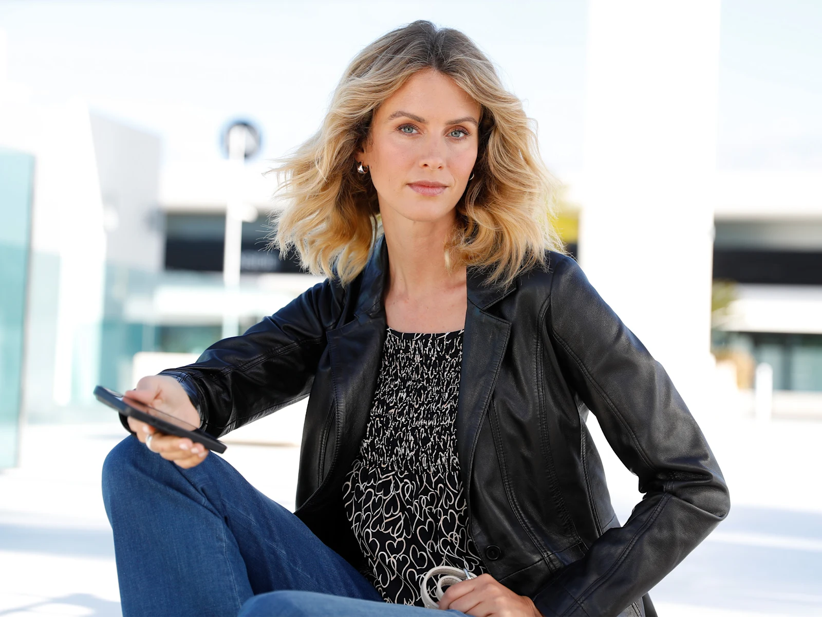 Ein Model sitzt draußen, trägt eine schwarze Lederjacke, ein gemustertes Oberteil und blaue Jeans, hält ein Smartphone. Ein Model sitzt draußen, trägt eine schwarze Lederjacke, ein gemustertes Oberteil und blaue Jeans, hält ein Smartphone.