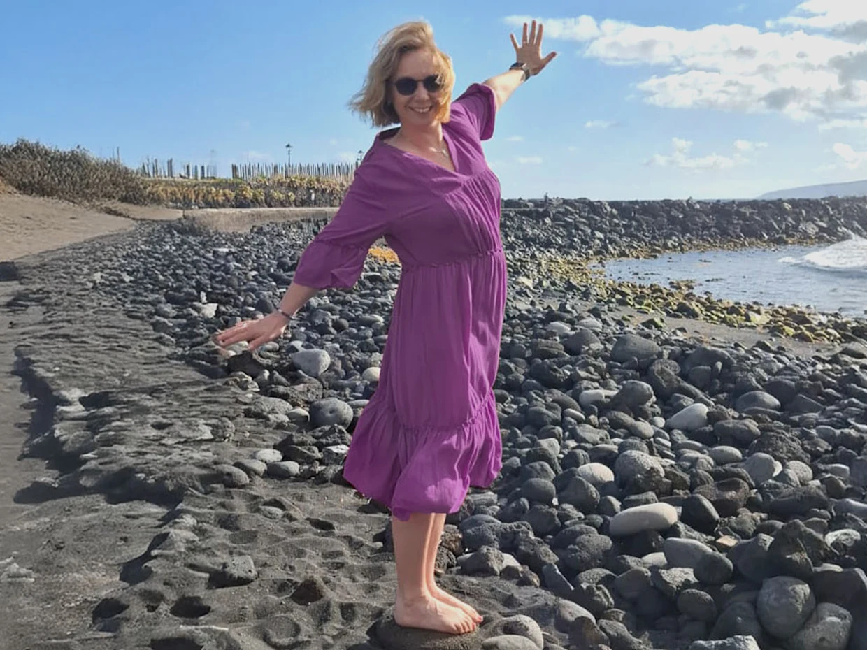 Eine Frau steht barfuß auf einem Kiesstrand, trägt ein lila Kleid und breitet die Arme aus. Eine Frau steht barfuß auf einem Kiesstrand, trägt ein lila Kleid und breitet die Arme aus.