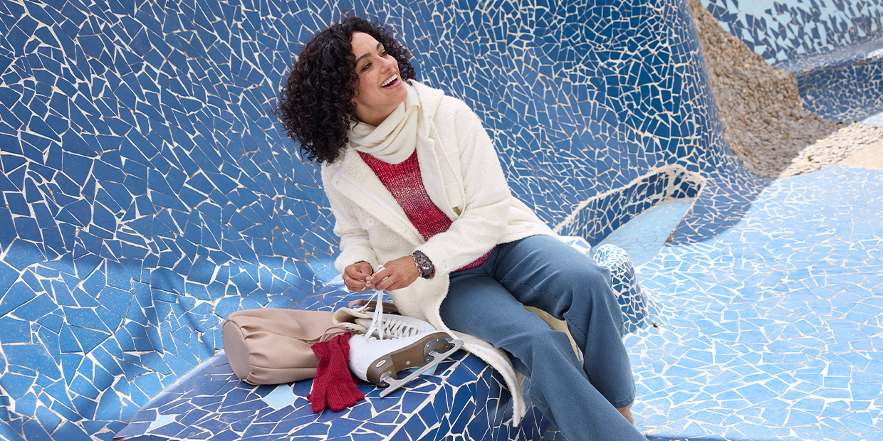 Ein Model sitzt auf einer blauen Mosaikbank, trägt einen ecrufarbenen Mantel, roten Pullover, eine blaue Hose und einen weißen Schal, neben ihr liegen eine Tasche und Handschuhe.