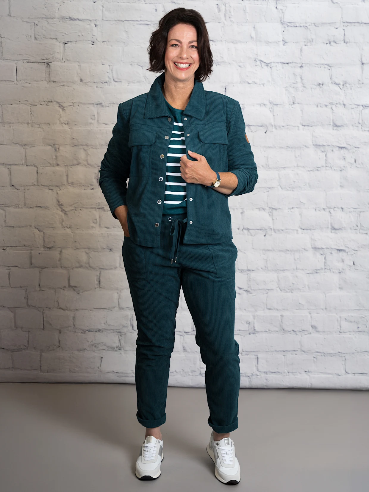 Eine dunkelhaarige Frau trägt eine petrolfarbene Cordjacke mit Hemdkragen, ein petrolfarbenes Ringelshirt mit Rundhals-Ausschnitt, eine petrolfarbene Cordhose mit Rundum-Dehnbund und weiß-silberfarbene Sneakers. Der Hintergrund ist eine weiße Wand.