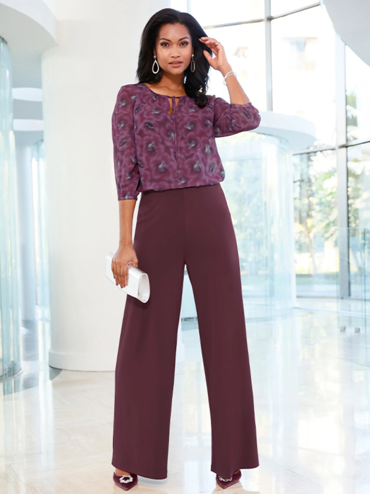 Ein Model trägt eine gemusterte, violette Bluse mit 3/4-Ärmeln, weite, dunkelviolette Hosen, passende Schuhe und eine weiße Clutch. Ein Model trägt eine gemusterte, violette Bluse mit 3/4-Ärmeln, weite, dunkelviolette Hosen, passende Schuhe und eine weiße Clutch.