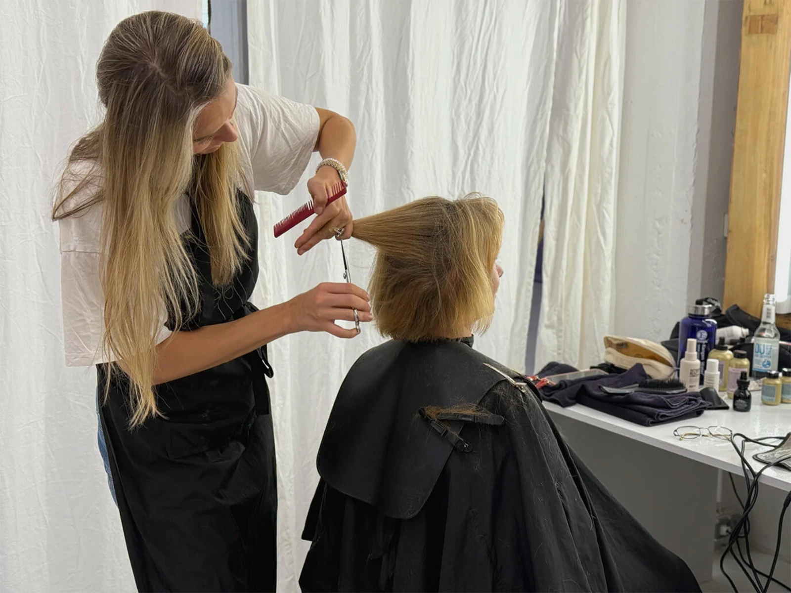 Eine Frau sitzt mit dem Rücken zur Kamera auf einem Stuhl und trägt einen schwarzen Friseurumhang. Sie erhält einen Haarschnitt von einer Friseurin, die mit einer Schere und einem Kamm arbeitet. Eine Frau sitzt mit dem Rücken zur Kamera auf einem Stuhl und trägt einen schwarzen Friseurumhang. Sie erhält einen Haarschnitt von einer Friseurin, die mit einer Schere und einem Kamm arbeitet.