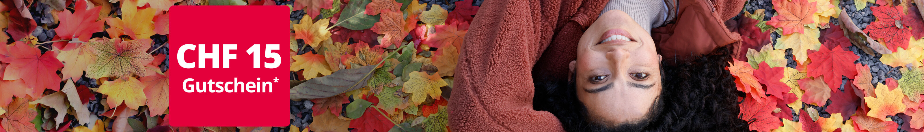 Eine junge, lächelnde Frau liegt auf dem Rücken in buntem Herbstlaub. Sie trägt eine kuschelige, rostbraune Fleece-Jacke. Links oben im Bild ist ein rotes Feld mit der Aufschrift 'CHF 15 Gutschein*' zu sehen.