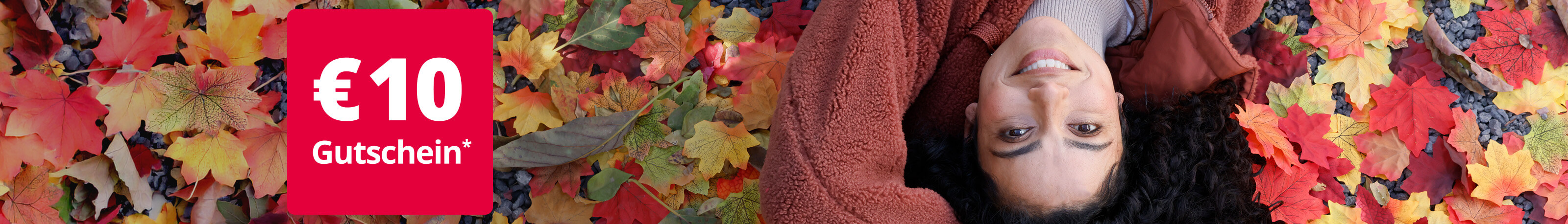 Eine junge, lächelnde Frau liegt auf dem Rücken in buntem Herbstlaub. Sie trägt eine kuschelige, rostbraune Fleece-Jacke. Links oben im Bild ist ein rotes Feld mit der Aufschrift '€ 10 Gutschein*' zu sehen.