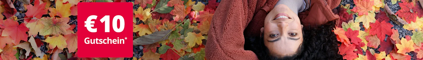 Eine junge, lächelnde Frau liegt auf dem Rücken in buntem Herbstlaub. Sie trägt eine kuschelige, rostbraune Fleece-Jacke. Links oben im Bild ist ein rotes Feld mit der Aufschrift '€ 10 Gutschein*' zu sehen.