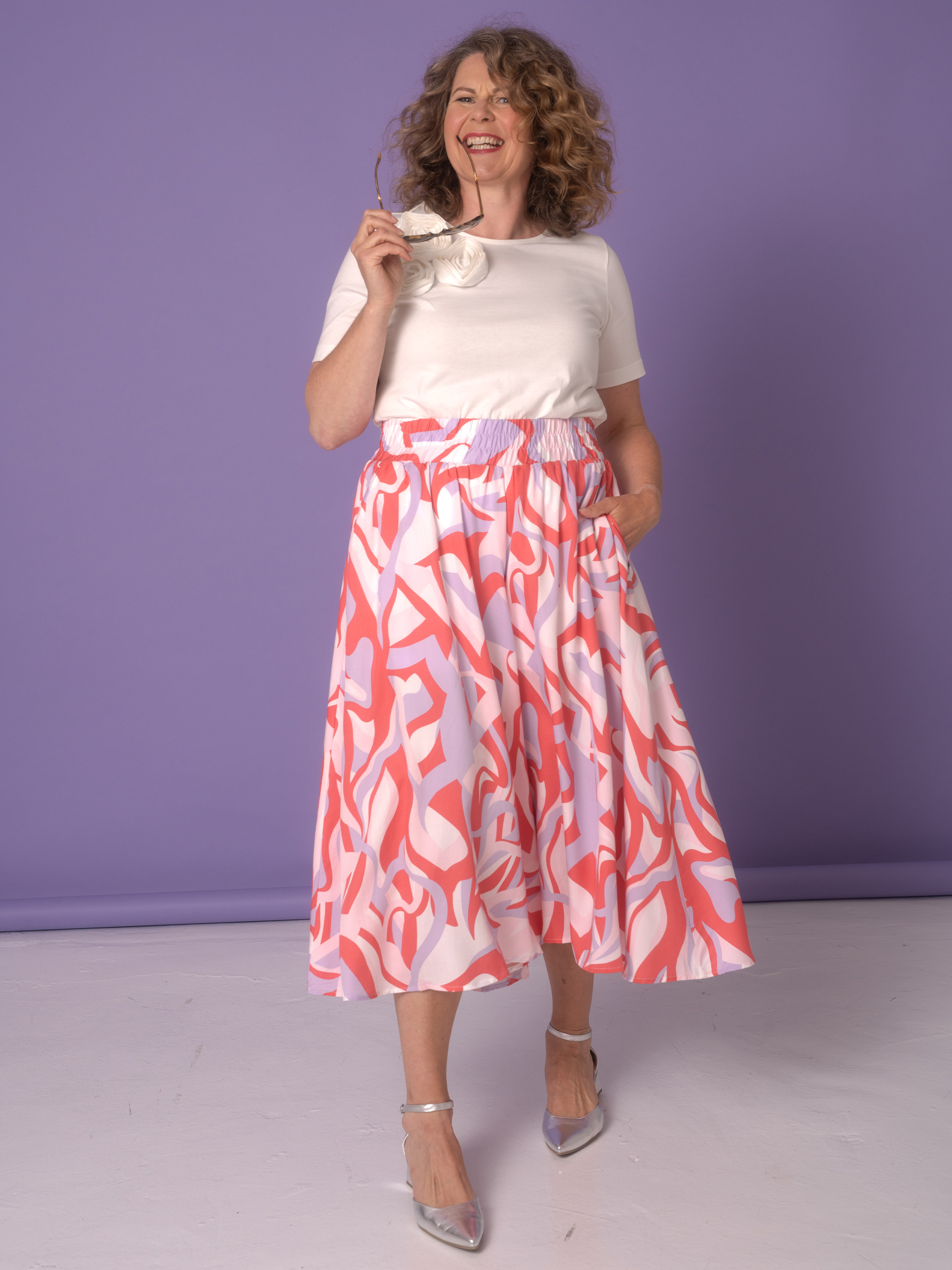 Eine Frau trägt ein weißes Oberteil und einen langen, gemusterten Rock in Rosa- und Lilatönen. Sie hält eine Brille in der Hand.